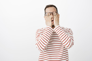Portrait of exhausted uncomfortable male model in striped pullover and glasses, rubbing eyes,...