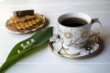 Lily of the valley, a cup of coffee and dessert on a white wooden table.