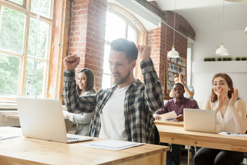 Happy successful worker excited with good results on stock market, raising hands up with yes gesture, colleagues congratulating with achievement. Trader celebrating online win in coworking office.