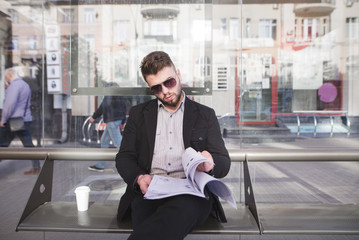 An office worker in a suit sitting at a bus stop and reading documents. A stylish businessman in...