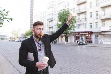 Businessman is standing by the road, eating a sandwich, drinking coffee, and catching a taxi. Busy...