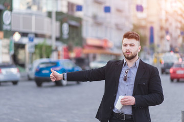 A solid bearded man in a business suit stands by the road with a cup of coffee in his hands and catches a taxi. Busy business man catches the car on the road