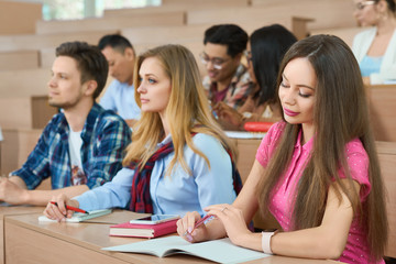 Fototapeta na wymiar Students sitting on wooden desks in modern university classroom. Boys and girls wearing colorful casual clothes. Guys reading, studuing, learning, writing. Looking happy. Friendly groupmates.