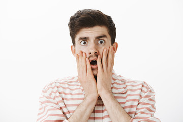 Studio shot of shocked and anxious handsome male model with moustache, touching cheeks with hands...
