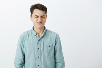 Waist-up portrait of disgusted european male coworker in shirt, smelling stinky smell and frowning, showing antipathy with grimace, standing against gray background disappointed