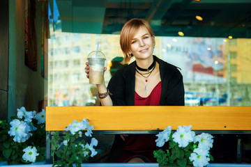 A smiling hipster girl with coffee-raf sits on a summer street cafe.