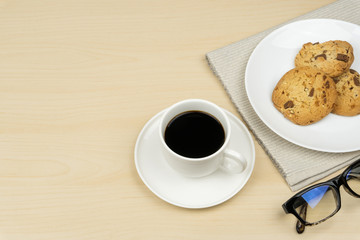 a cup of coffee, three pieces of chocolate chip cookies in a white round dish and the eyeglasses on the wooden table.