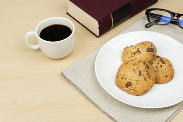 a cup of coffee, three pieces of chocolate chip cookies in a white round dish, a red book and an eyeglasses on the wooden table.