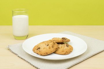 a glass of milk, three pieces of chocolate chip cookies in a white round dish on the wooden table. green background