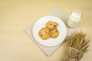 a glass of milk, three pieces of chocolate chip cookies in a white round dish and dried wheat bouquet on the wooden table.