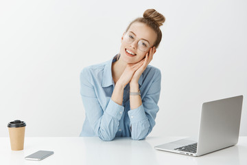 Dreamy good-looking european female in stylish glasses sitting in office near laptop and smartphone, drinking coffee, working on project, smiling broadly at camera, imaging vacation with boyfriend