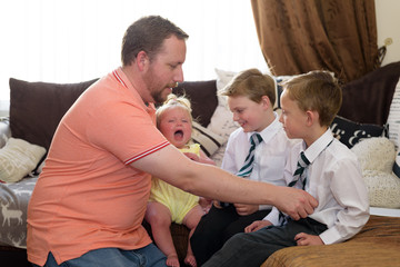 Father getting two sons dressed for school at home whilst holding baby girl in arms