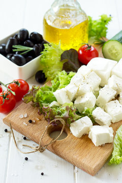 Cooking qreek salad with fresh vegetables, feta cheese and black olives on a white wooden table.