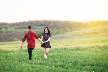 Beautiful couple holding their hands walking at the green blooming field at sunset. Woman looking at the camera. Outdoor