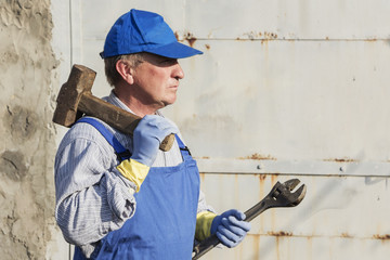 worker in blue overalls with a sledgehammer and a wrench. Serious look.