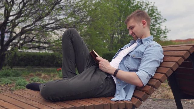 The young man sits in the park and reads the book. The happy guy sits on a wooden bench and reads the book.