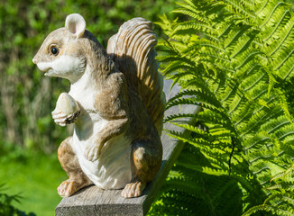 Decorative garden squirrel that holds one acorn stands on the wood handrail close to the leaves of fern , sunny day, like alive
