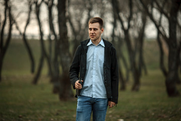 Vape teenager. Handsome young white guy in black jacket and checkered shirt vaping an electronic cigarette among the trees in the park at sunset. Lifestyle.