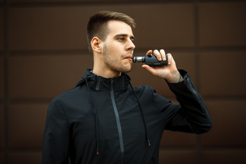 Vape teenager. Handsome young white guy in black jacket vaping an electronic cigarette opposite the futuristic urban background in the spring. Lifestyle.