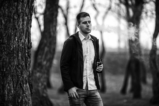 Vape teenager. Handsome young white guy in black jacket and checkered shirt vaping an electronic cigarette among the trees in the park in the spring. Black and white.