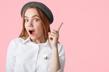 Surprised beautiful European female keeps fore finger raised, wears elegant shirt and hat, gets good idea to put in life, isolated over pink background with copy space for your promotional content