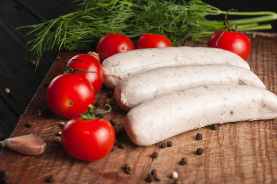 Three raw sausages on a wooden brown board on a black background with cherry tomatoes, parsley and dill, pepper and garlic.