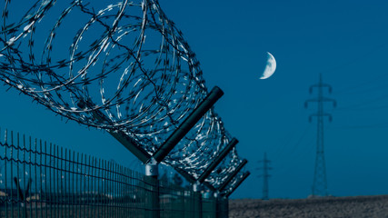 Close-up of sharp razor wire on fence. Gloomy blue sky and moon in background. Abstract industrial or military compound with barbed tape in a field with power line. Illuminated by artificial lighting.