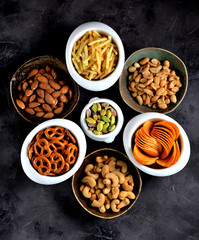 Different kinds of snacks - chips, salted peanuts, cashews, almonds and pistachios, pretzels with salt, potatoes, salted straw.