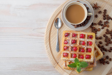 A pile of Belgian waffles with cranberries on a light cutting board and cup of coffee with milk and scattered coffee beans. Top view.