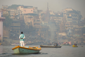 A man is sailing on his wooden boat in the sacred river Ganges in the city of Varanasi at sunset. Blurred Manikarnika ghat on background.