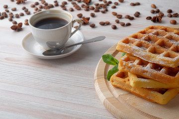 A pile of Belgian waffles on a light cutting board and cup of black coffee and scattered coffee beans. Copy space for text.