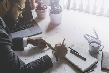 Bearded young businessman working at modern studio. Man sitting at wooden table using laptop and making notes. Blurred background.Visual effects