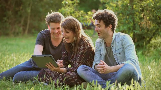 happy and smiling friends sitting on a meadow using Digital Tablet
