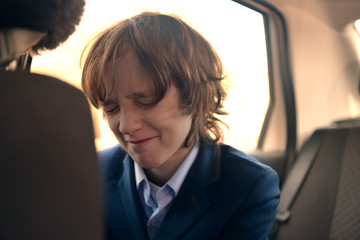 A boy is a teenager with a long hair in a classic suit in the car. Stylish child in the car.