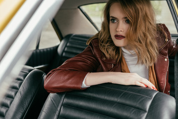 Obraz na płótnie Canvas beautiful young woman in leather jacket sitting in classic car and looking away