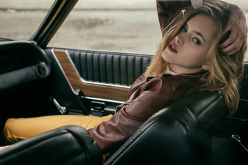 Fototapeta na wymiar seductive young woman in leather jacket looking at camera while sitting in car