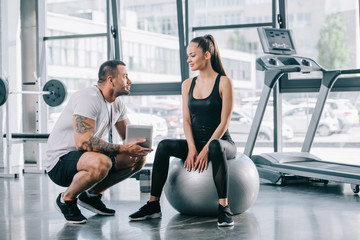 young sportswoman sitting on fitness ball and talking to personal trainer with digital tablet at gym