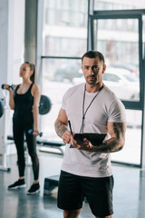 personal trainer with clipboard and young sportswoman exercising with dumbbells behind at gym