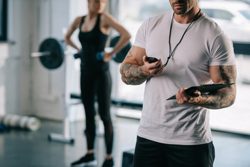 cropped shot of male personal trainer looking at timer and young sportswoman exercising with dumbbells behind at gym