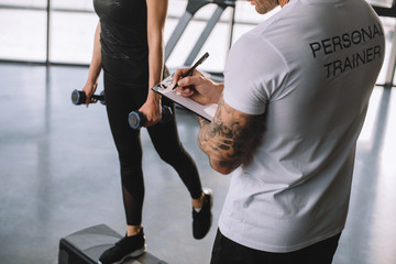 cropped image of male personal trainer writing in clipboard and young sportswoman doing step aerobics exercise with dumbbells at gym - obrazy, fototapety, plakaty