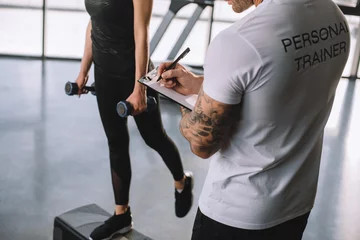 Poster cropped image of male personal trainer writing in clipboard and young sportswoman doing step aerobics exercise with dumbbells at gym © LIGHTFIELD STUDIOS