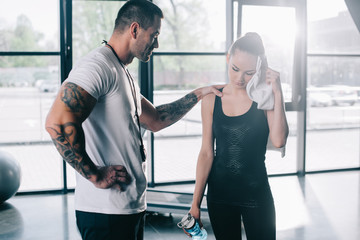 Fototapeta na wymiar male personal trainer cheering up young sportswoman with bottle of water wiping face with towel at gym