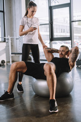 female personal trainer using timer while sportsman doing exercises on fitness ball at gym