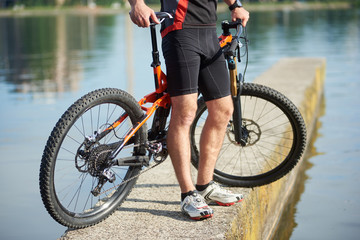 Crop photo of sporty male cyclist wearing professional sportswear standing near bicycle on pier next to city lake. Concept of healthy lifestyle, outdoor training