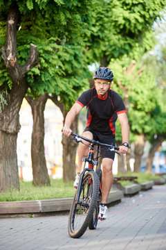 Sporty man bicyclist in professional cycling clothing and protective helmet riding on bicycle along empty city streets, green trees around. Sportsman performing morning outdoor exercise and training.
