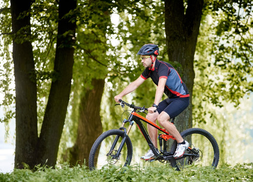 Sporty man cyclist in cycling clothing and protective helmet riding on bicycle through sunny alley among trees in green beautiful park. Concept of healthy lifestyle, exercising, fresh air