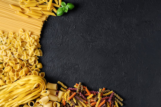 Raw Italian pasta fettuccine, paccheri, farfalle, spaghetti, fusilli, penne, conchiglie on a dark background.