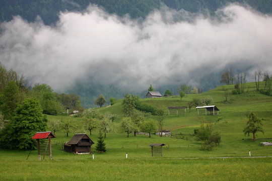Krajobraz z zielonymi łagodnymi pagórkami, chatami drewnianymi, gęstą opadającą z góry mgłą