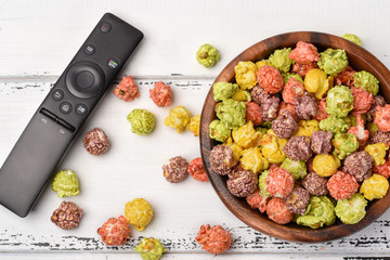 Color caramel popcorn in a wooden bowl on a white wooden background with remote control for TV