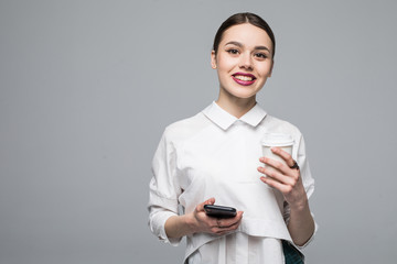 Portrait of a cheery young girl using mobile phone while holding coffee cup isolated over white background